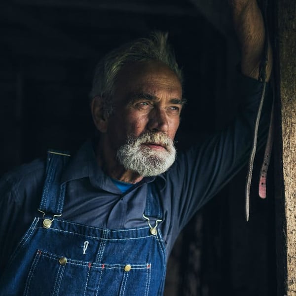 Dirk Volcano with white beard wearing blue denim overalls and a dark shirt, leaning against a wooden beam in a rustic setting with a thoughtful expression.